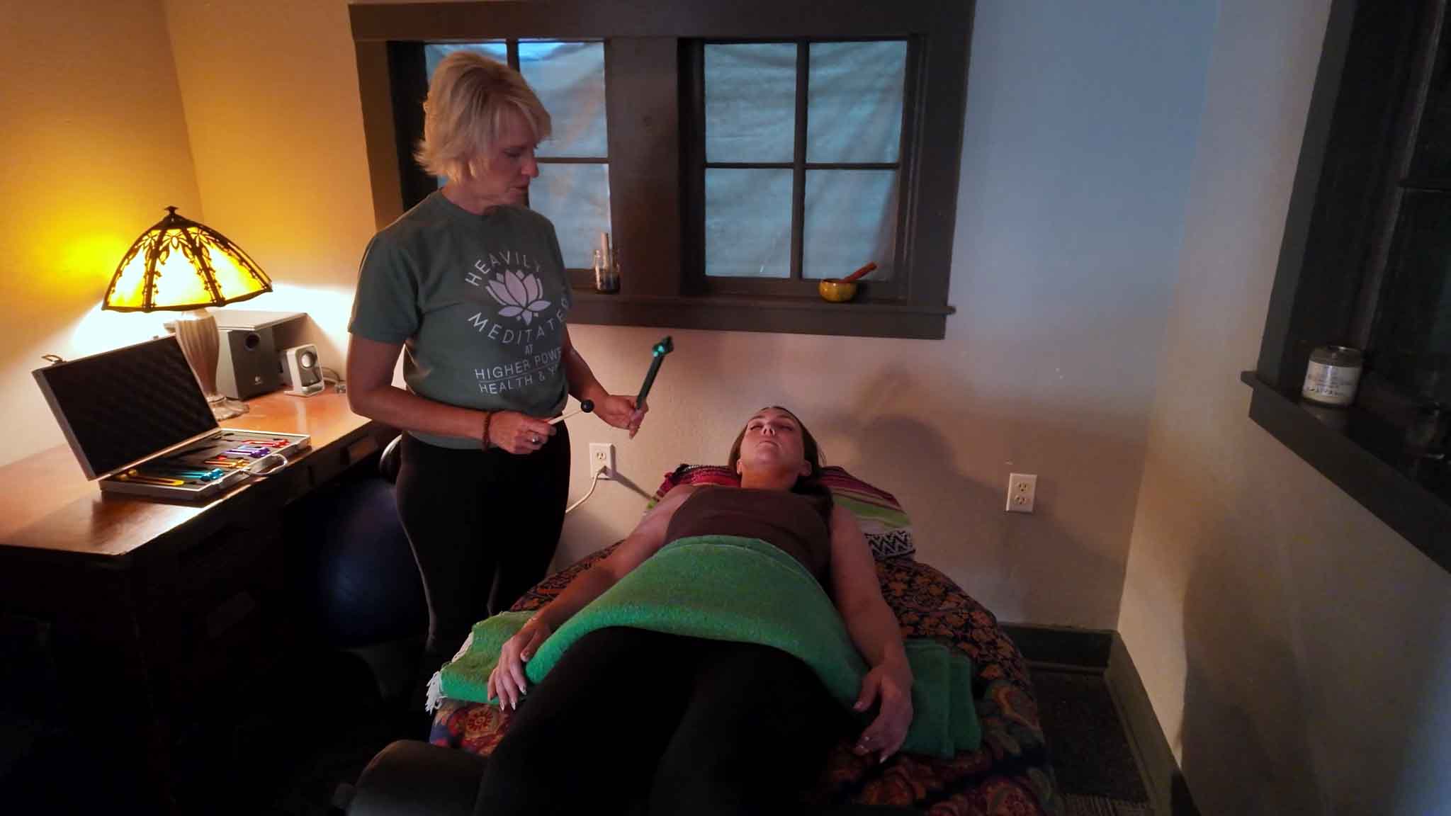 Michelle Robert holding a weighted tuning fork above a client who is resting, fully clothed and covered with a blanket, on the treatment table.